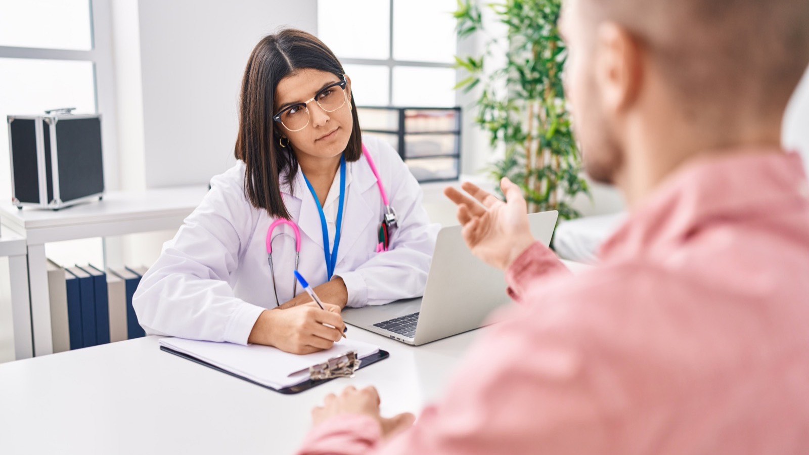 Paciente tira dúvidas com a médica na consulta enquanto ela anota na prancheta, com o notebook aberto sobre a mesa