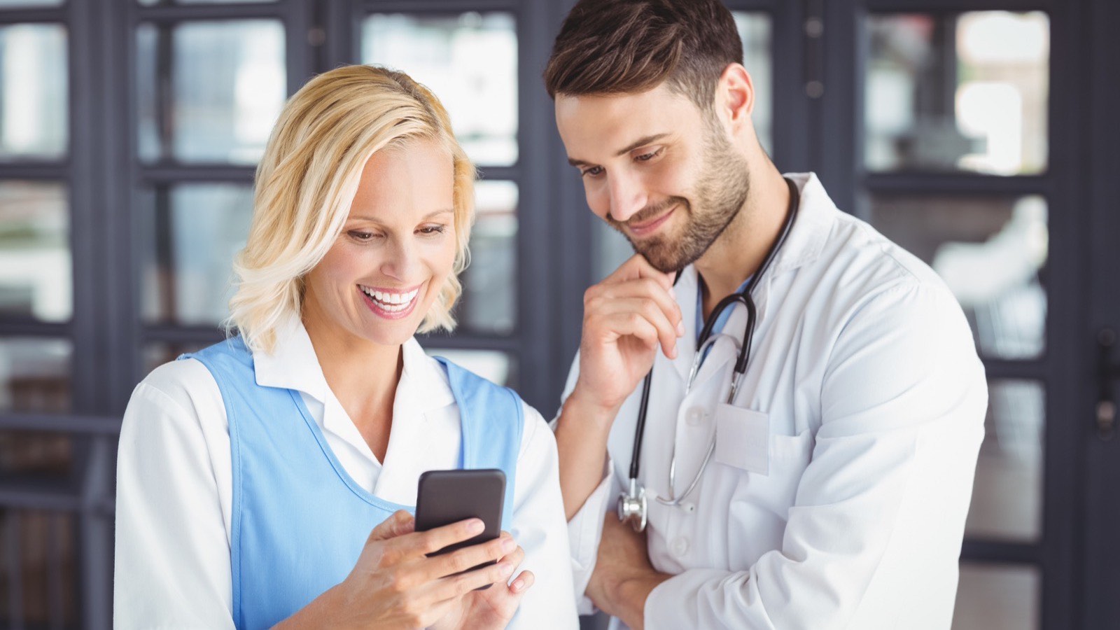 Médico e assistente da clínica conferem juntos uma mensagem no celular durante a rotina do consultório