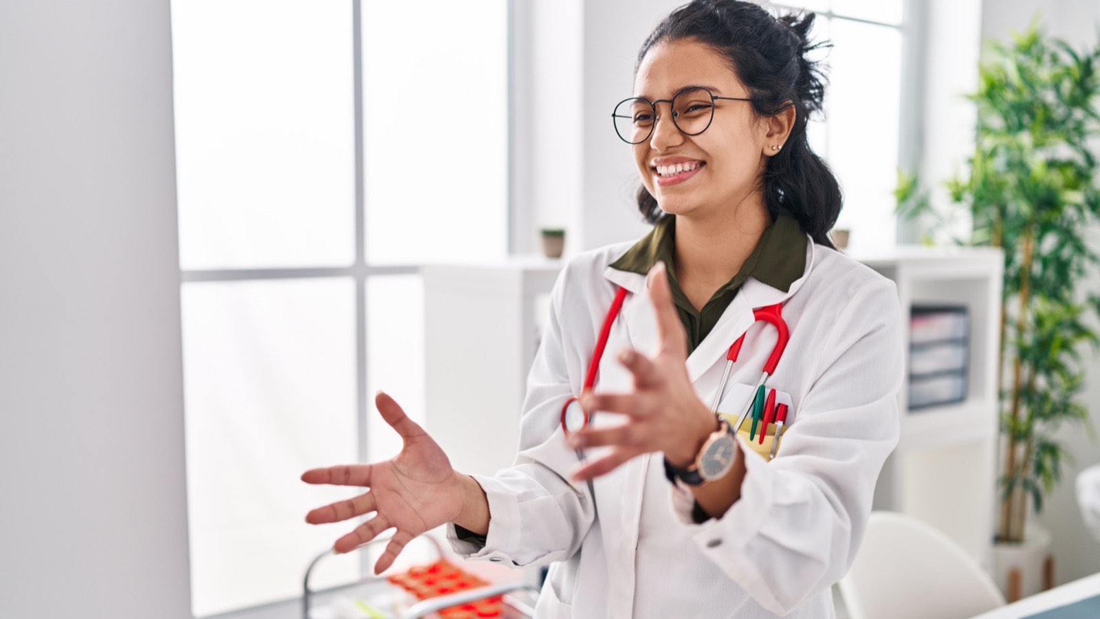 Médica de óculos e jaleco sorri enquanto conta sua experiência, em pé no consultório claro