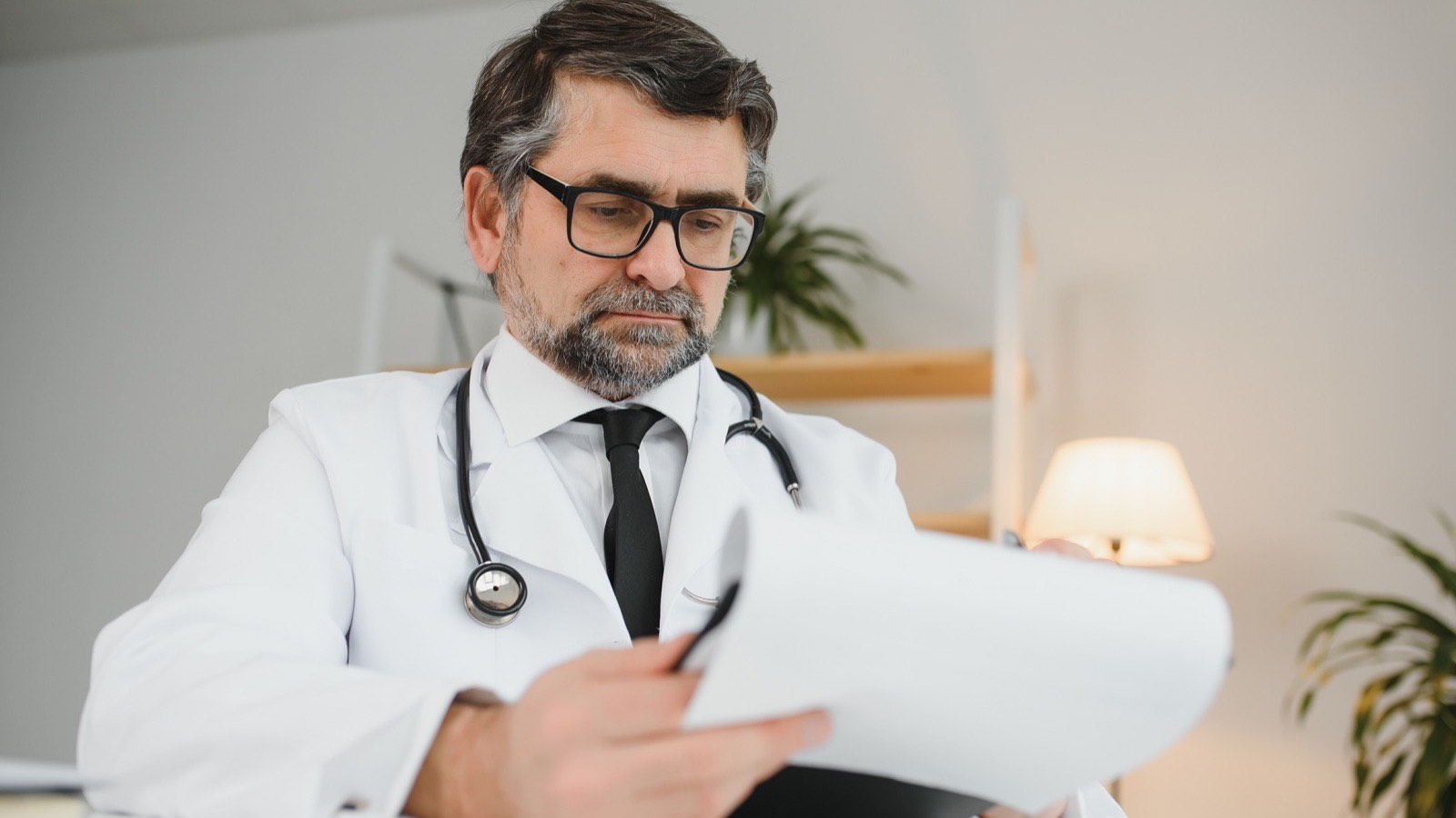 Médico de jaleco e óculos revisando com atenção um texto impresso na mesa do consultório