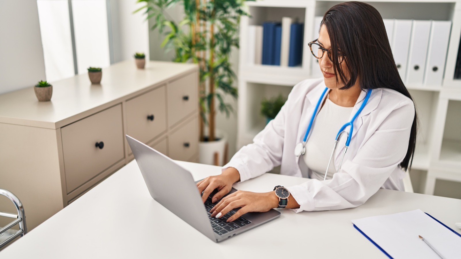 Médica de jaleco e óculos digitando no notebook na mesa do consultório, com uma prancheta ao lado