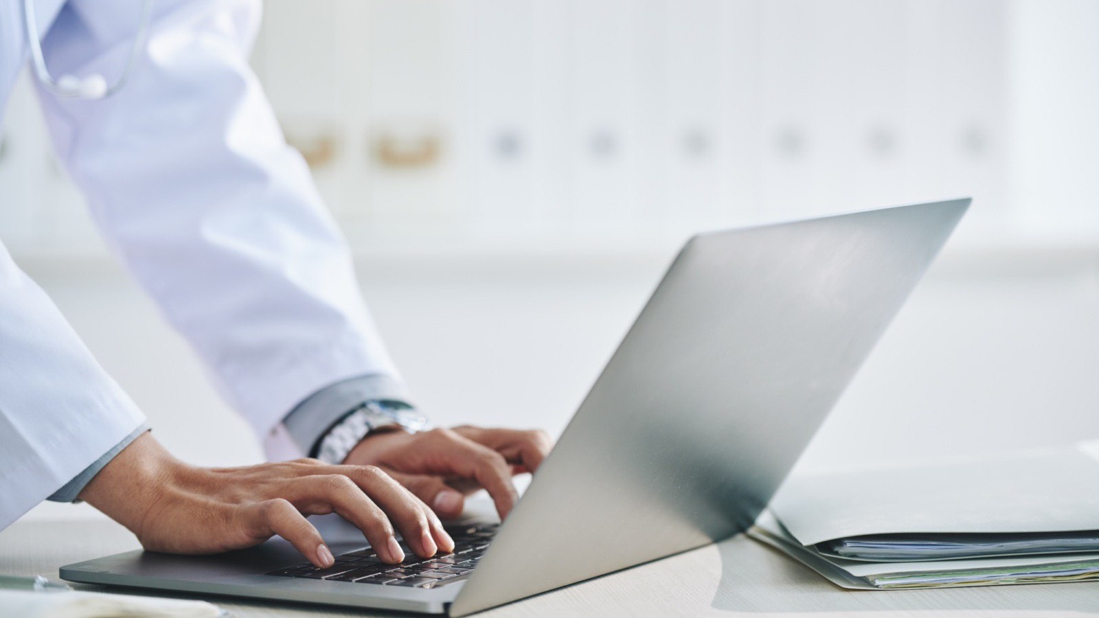 Mãos de médica de jaleco digitando no notebook sobre a mesa do consultório, ao lado de pastas de documentos