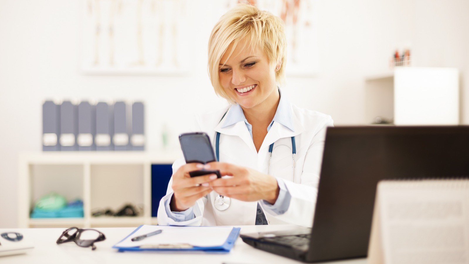 Médica sentada à mesa do consultório lê uma mensagem no celular, com o notebook aberto ao lado