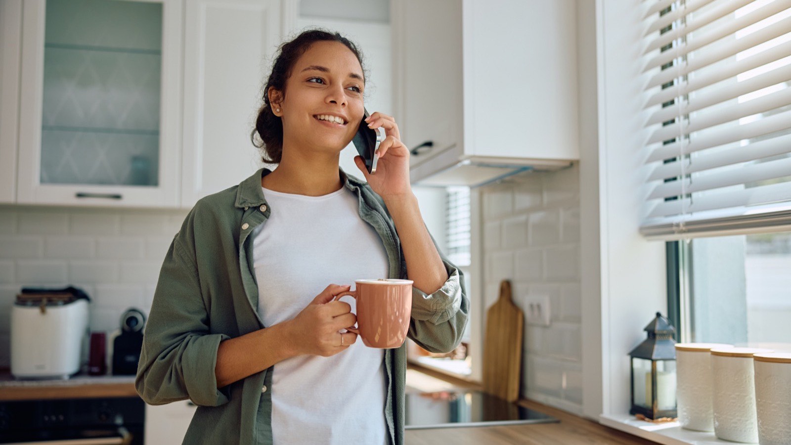 Mulher na cozinha de casa ligando pelo celular para marcar consulta, com uma caneca de café na mão