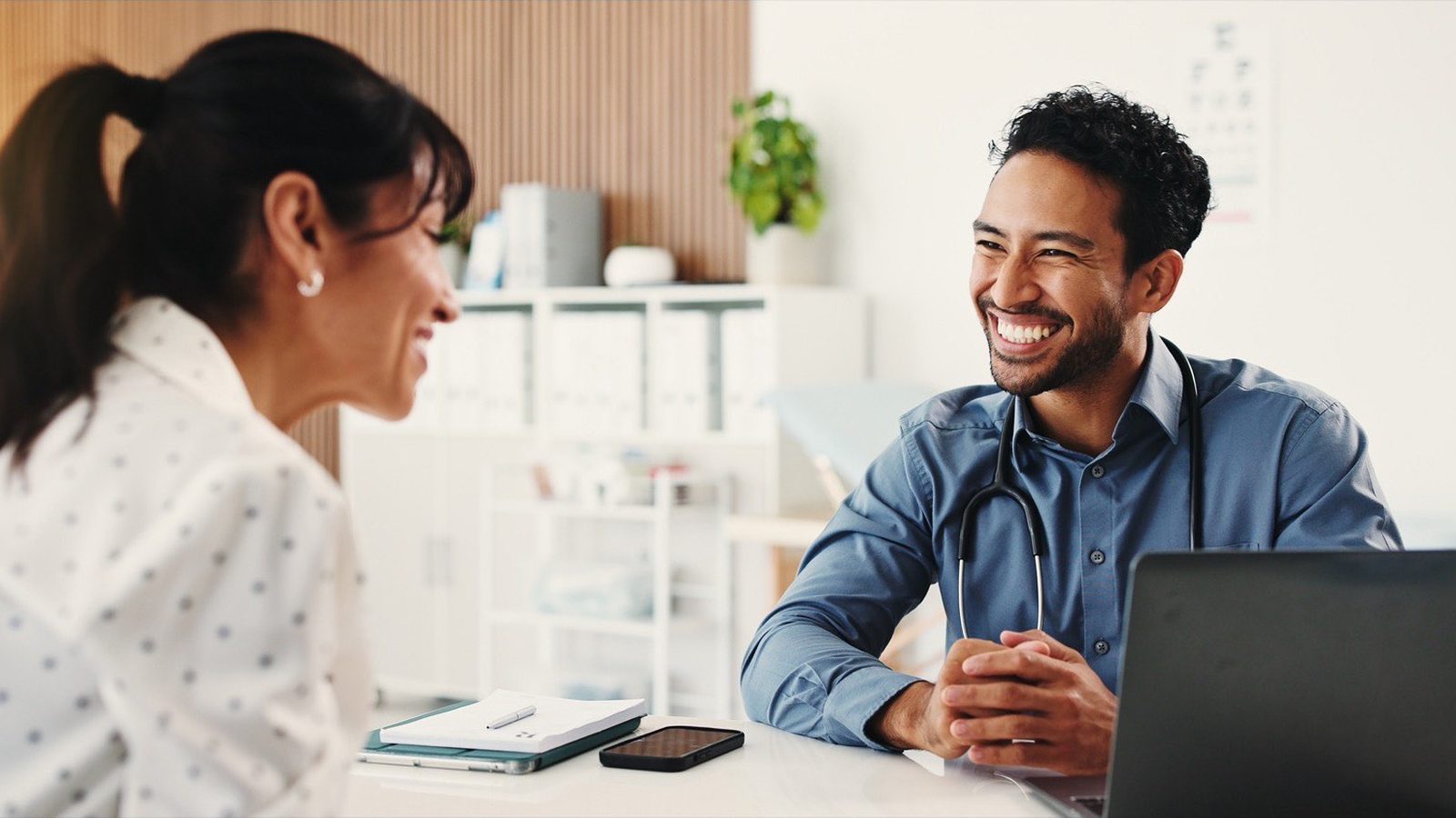 Médico de estetoscópio conversa sorrindo com uma profissional de marketing diante do notebook no consultório